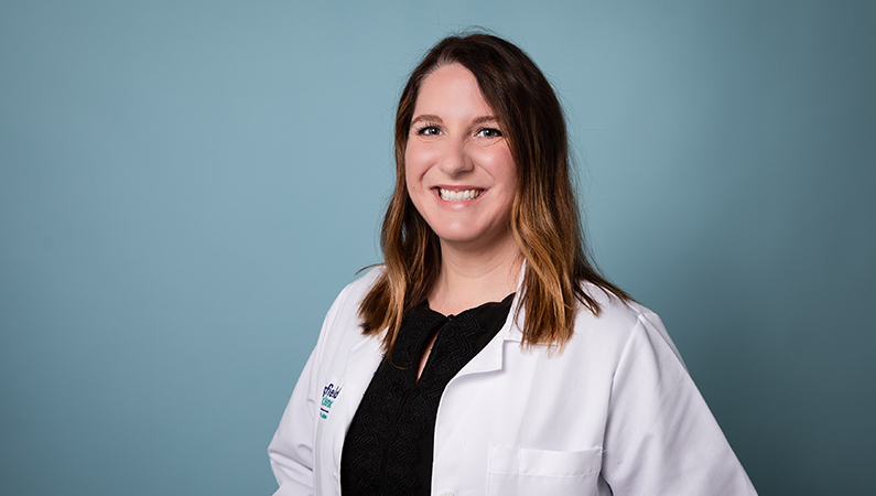 Female nurse practitioner in white coat smiling in front of a light blue backdrop.