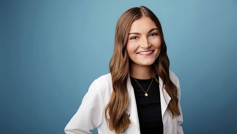 Female wearing white medical coat smiling in front of blue background