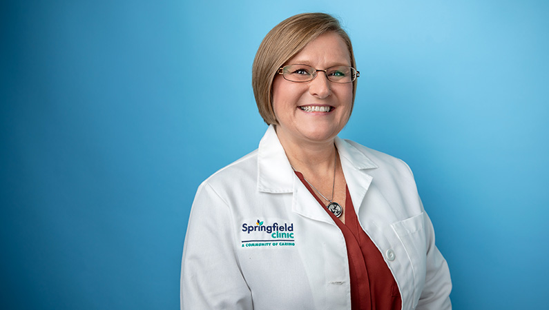 Female nurse practitioner in white medical coat smiling in front of a light blue photo backdrop.
