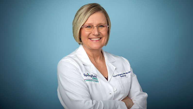 Female nurse practitioner in white medical coat smiling in front of a light blue photo backdrop.