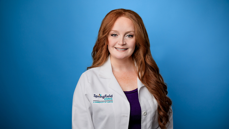 Female with long red hair wearing white medical coat smiling in front of light blue backdrop.