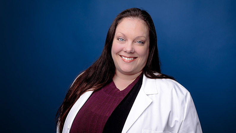 Female doctor in white medical coat smiling in front of royal blue photo backdrop.