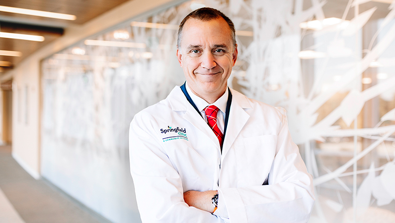 Male wearing white medical coat posing in well-lit walkway.