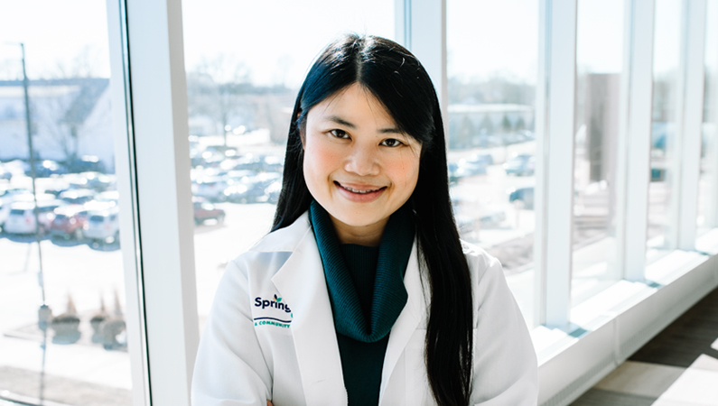 Female with dark hair wearing white medical coat smiling in front of windows