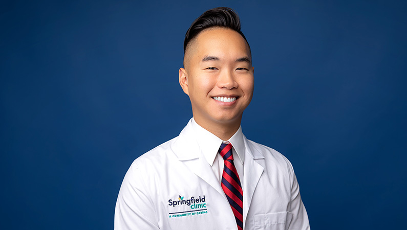 Male in white medical coat with arms crossed smiling at camera in front of light blue photo backdrop.
